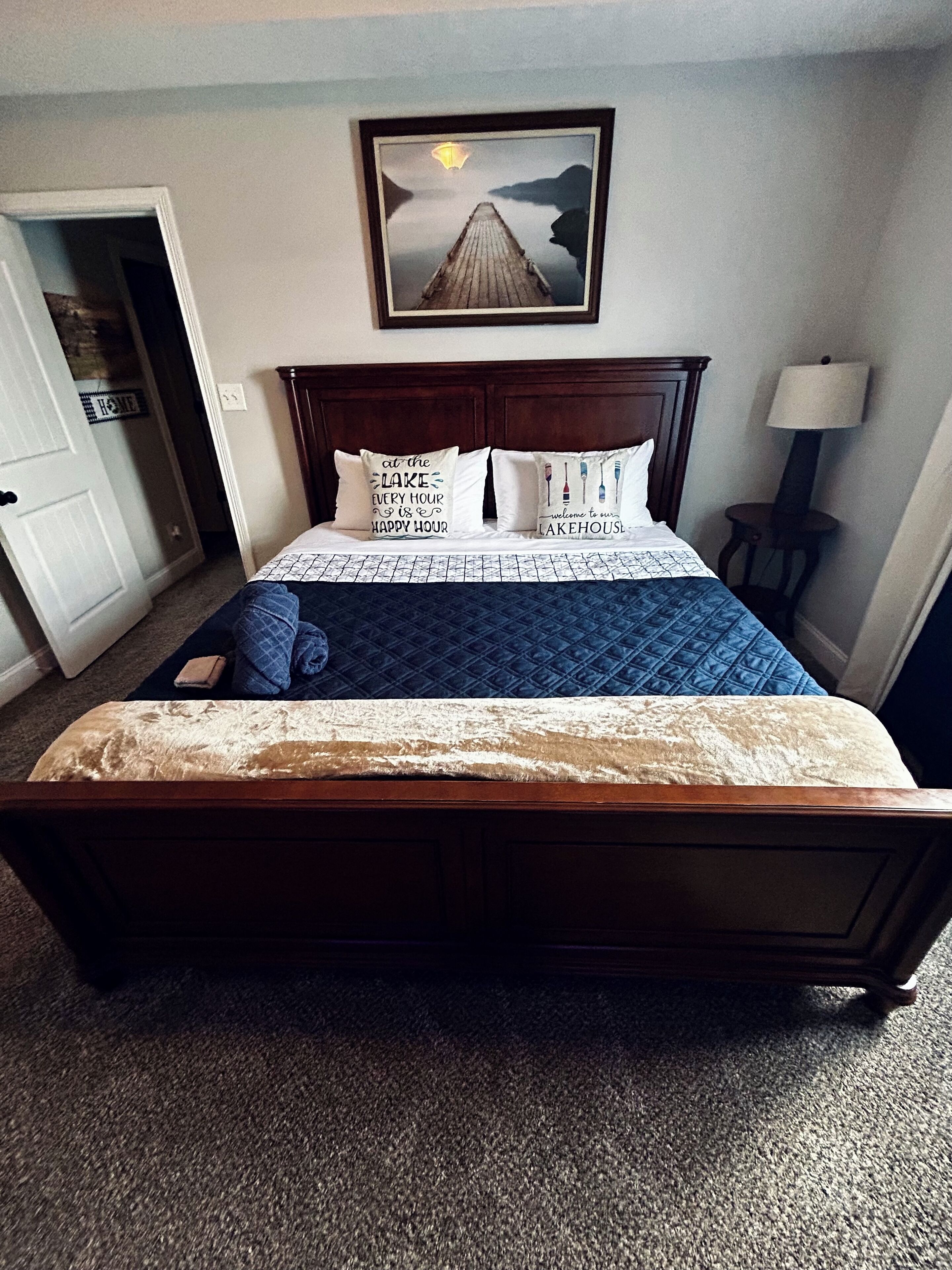 A hotel room with a wooden bed, a framed picture above the bed, a bedside lamp, and a door to the left.