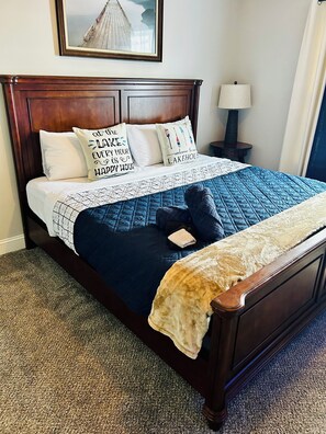A neatly made bed with a blue quilt, two pillows with text, a small bedside table with a lamp, and a framed picture on the wall.
