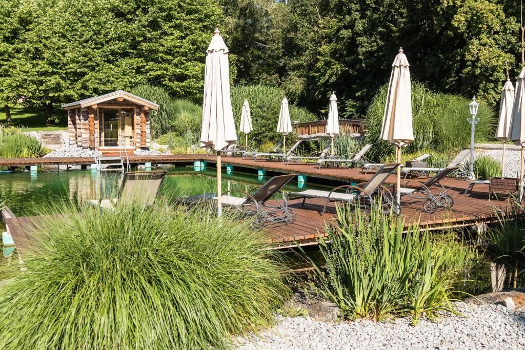 Una cabaña de madera con un muelle que se adentra en un lago tranquilo, rodeada de exuberante vegetación y equipada con sillones de descanso y sombrillas.
