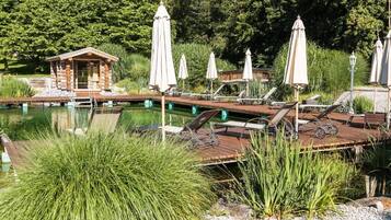 Una cabaña de madera con un muelle que se adentra en un lago tranquilo, rodeada de exuberante vegetación y equipada con sillones de descanso y sombrillas.
