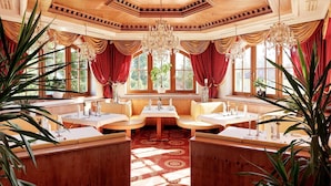 A restaurant with a high ceiling, wooden paneling, and large windows with red curtains. Tables are set with white tablecloths and candles. - Parkschlössl zu Thyrnau (Thyrnau)