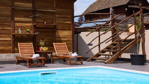 Un edificio de madera con piscina, dos sillas de madera, una mesa con un florero y fruta, y una escalera.