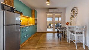 Una cocina moderna con gabinetes color turquesa, un refrigerador de acero inoxidable y una encimera de madera. Hay un comedor con sillas blancas y una mesa, además de un reloj decorativo en la pared.