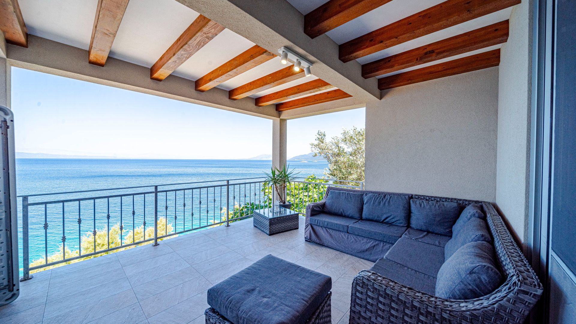 A covered outdoor seating area with a view of the sea, a sofa, an ottoman, and a potted plant.