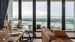 Un comedor con una mesa preparada para dos personas, un sofá y vistas a un río y al paisaje urbano a través de amplios ventanales.
