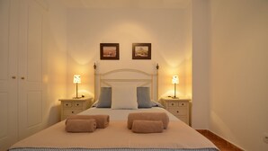 Una cama bien hecha con dos almohadas y dos toallas enrolladas. Hay dos mesitas de noche con lámparas y un cuadro enmarcado sobre la cama.