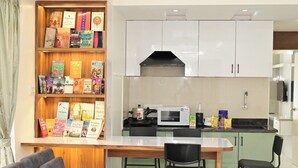 Una cocina compacta con microondas, estufa y diversos alimentos. Se observa una estantería llena de libros a la izquierda.