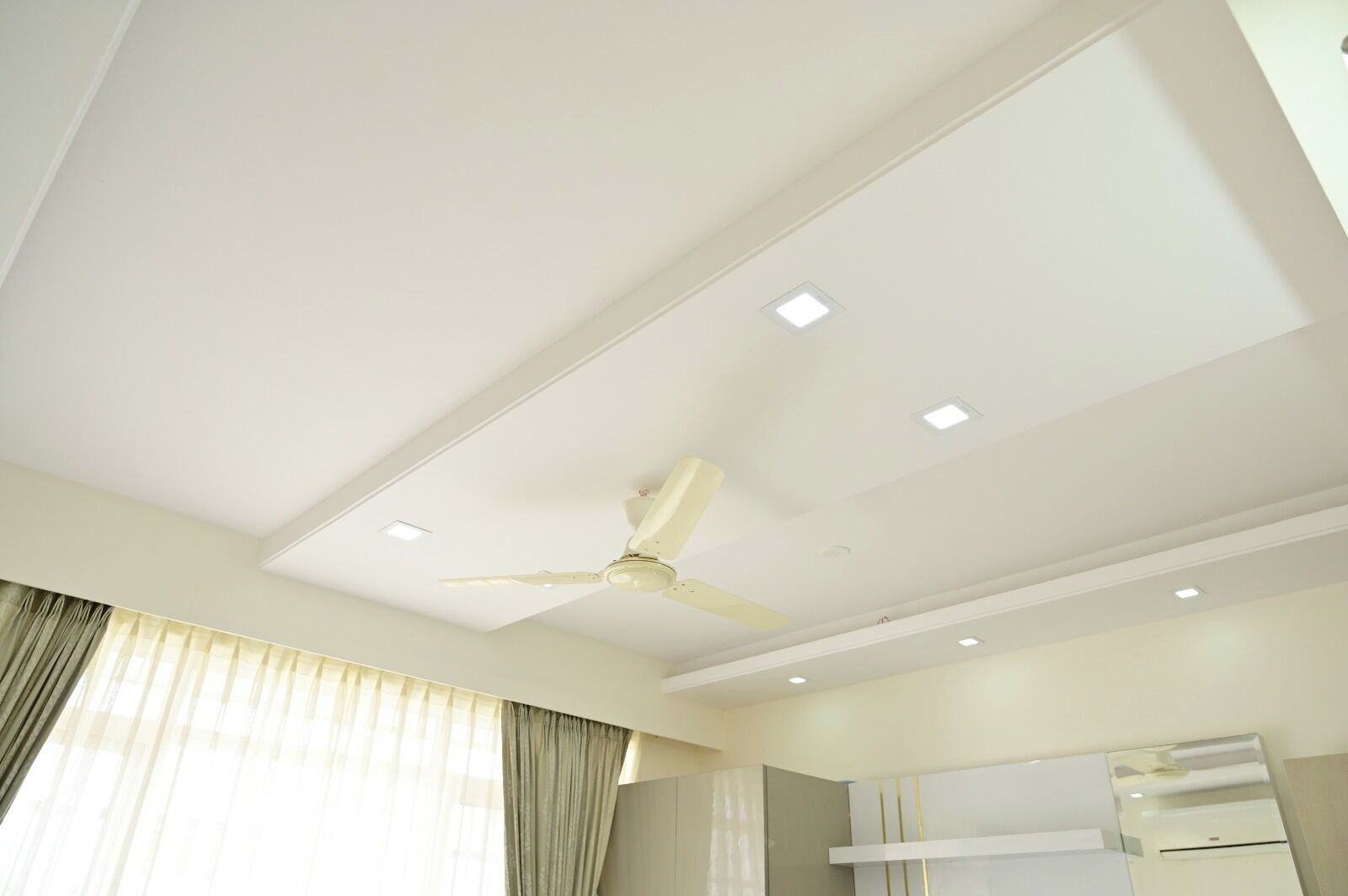 A room with a ceiling fan, recessed lighting, and a large window with curtains.