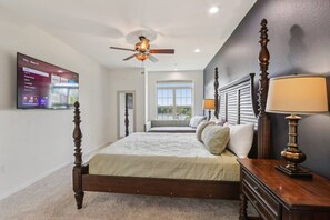 A bedroom with a four-poster bed, a ceiling fan, a television mounted on the wall, and a window with a view of the outdoors.
