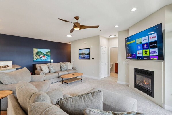 A modern living room with a large flat-screen TV displaying various streaming services, a ceiling fan, and a fireplace.