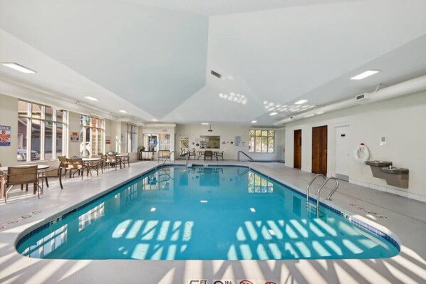 An indoor swimming pool with clear water, surrounded by tables and chairs, and a ceiling with recessed lighting. - Bushwood at Delton Grand (Wisconsin Dells)