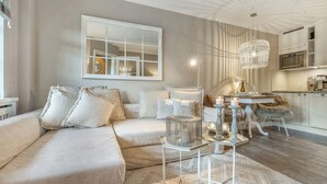 A modern living room with a beige sofa, a dining area with a white table, and a kitchen in the background. - Lütt'n Witthüs - Whg. 6 (Wenningstedt-Braderup (Sylt))