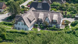 A large, white building with a steeply pitched roof, surrounded by lush greenery and a well-maintained garden. - Lütt'n Witthüs - Whg. 6 (Wenningstedt-Braderup (Sylt))