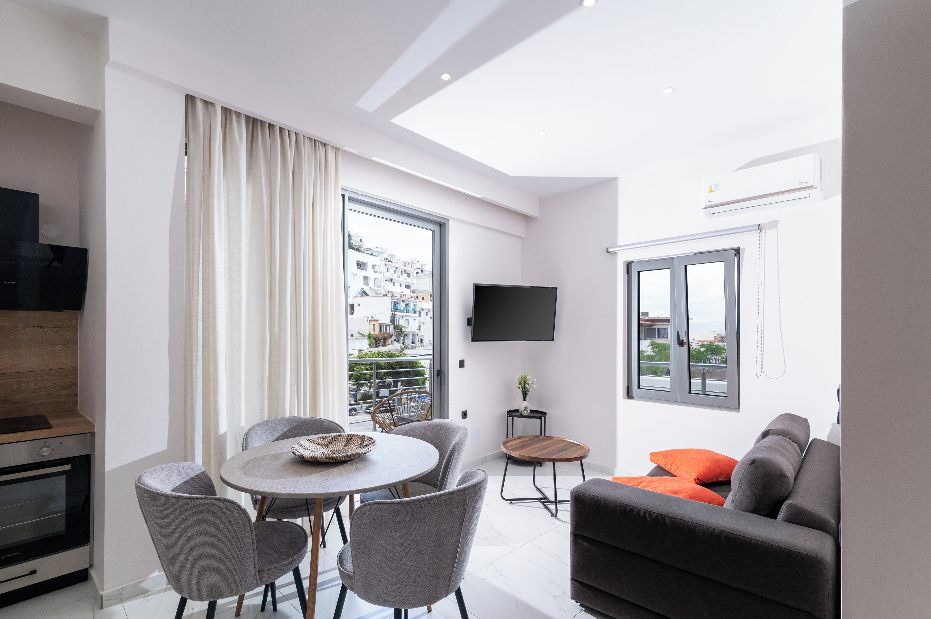 A modern living room with a dining area, a flat-screen TV mounted on the wall, and a balcony with a city view.