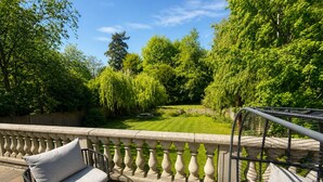 Une terrasse dotée d’une balustrade en pierre, d’un banc rembourré et offrant une vue sur un jardin bien entretenu, avec différents arbres et une pelouse.