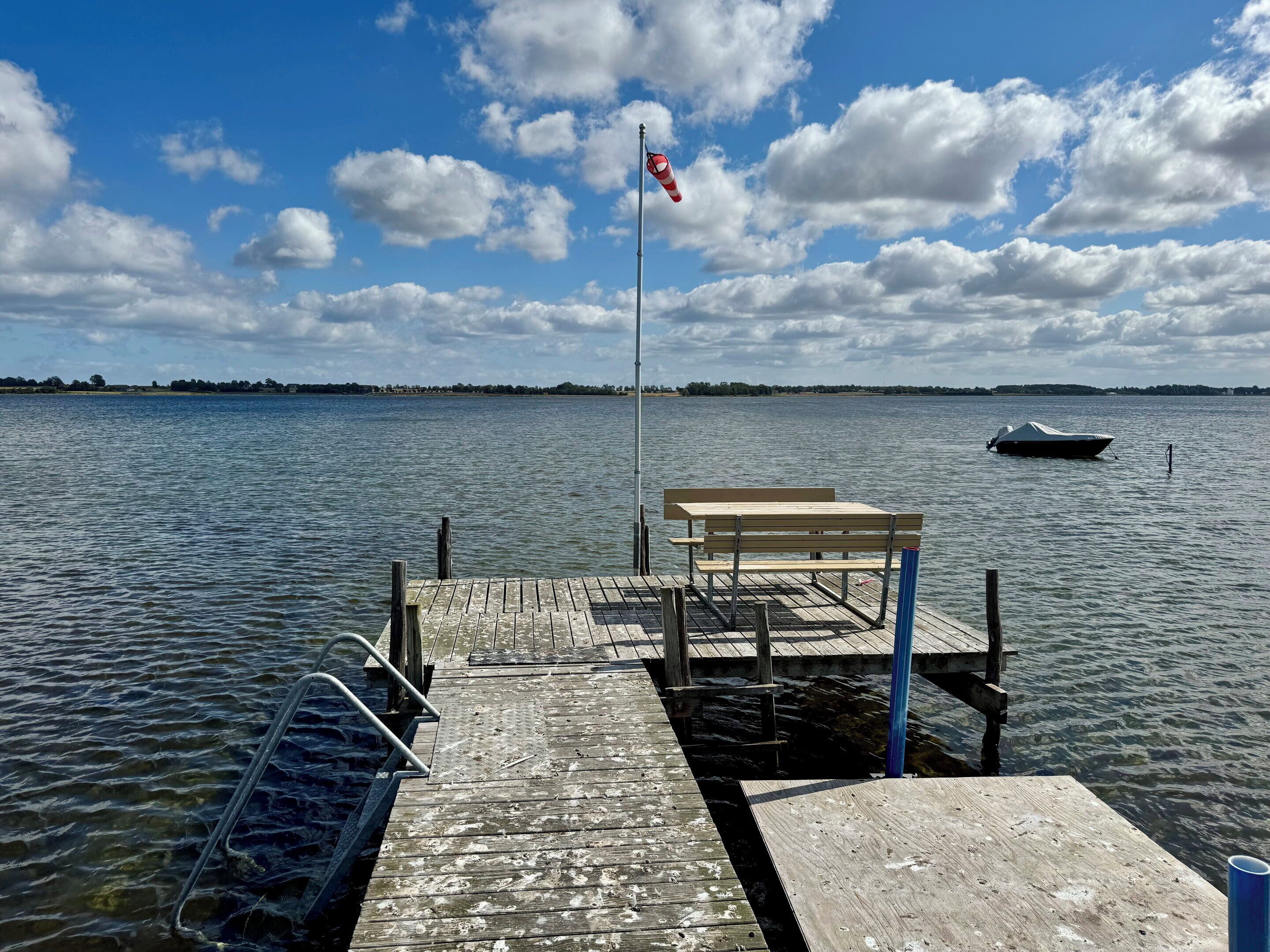 Une jetée en bois s’étendant sur une étendue d’eau calme, un banc, un mât avec un drapeau, et un bateau au loin.