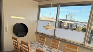 A dining area with a table, chairs, a hanging light fixture, and a view through large windows.