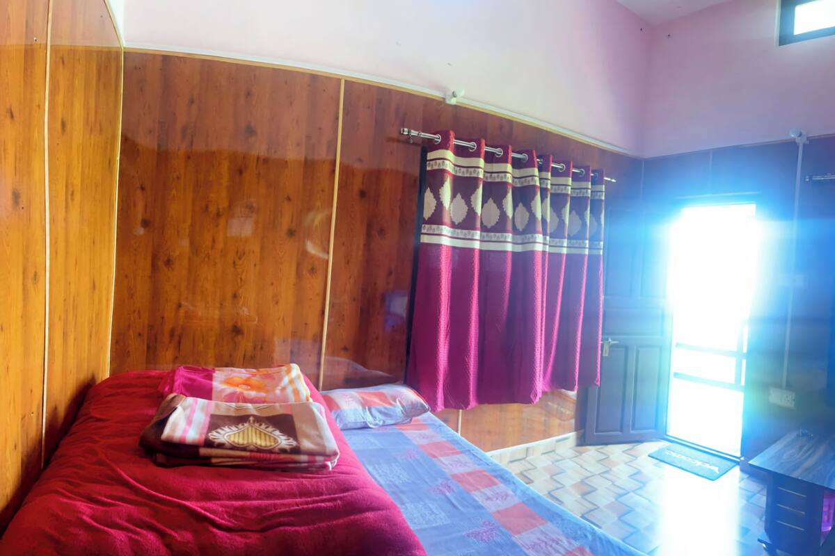 A room with wooden walls, a bed with a red blanket, a quilt, and a window with curtains.