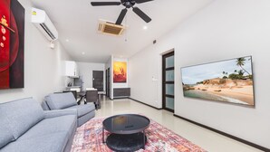Uma sala de estar moderna com um sofá cinzento, uma mesa de centro redonda e um tapete colorido. Há uma TV na parede exibindo uma cena de praia.