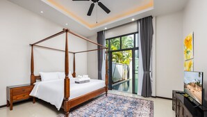 Uma cama de dossel com um edredom branco, uma mesa de cabeceira de madeira, um tapete estampado, uma televisão sobre um móvel de madeira e vista para uma piscina através de grandes janelas.