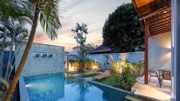 Piscina com piso cerâmico, cercada por palmeiras e uma parede de pedra.