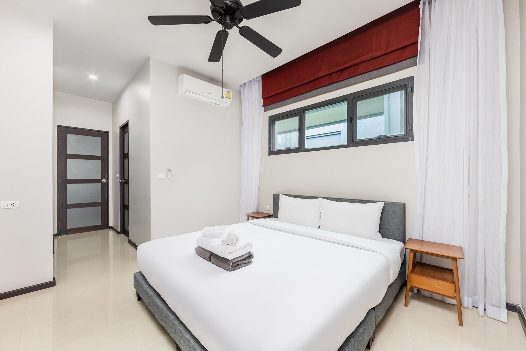 Una habitación de hotel moderna con una cama grande, un ventilador de techo, un equipo de aire acondicionado, una ventana con cortinas y una mesita de noche pequeña.