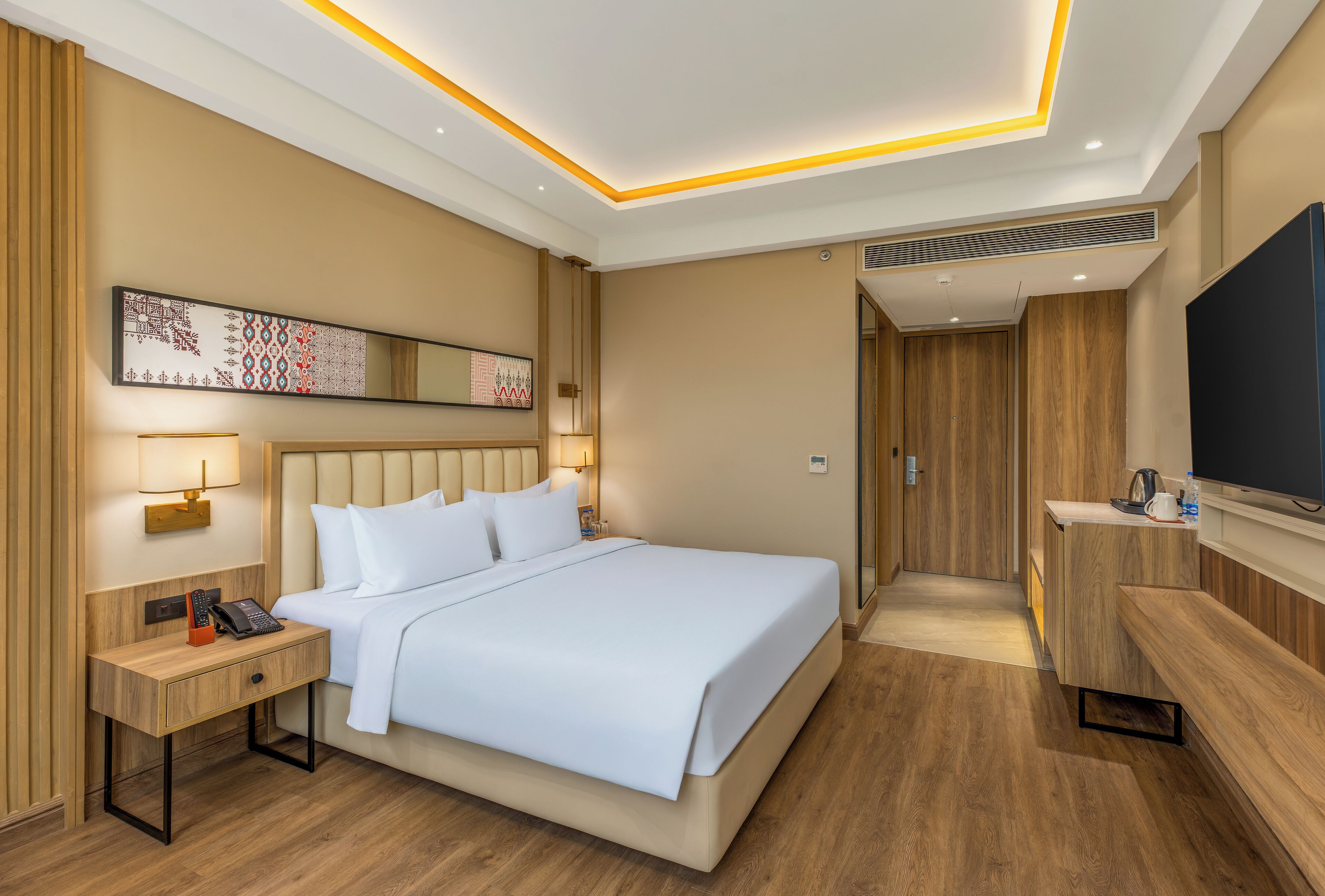A hotel room with a bed, bedside table, lamp, artwork, and a TV.