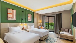 A hotel room with two beds, a sofa, a small table, and a view of greenery. - The Aaureum Resort & International Convention Centre (Bhilwara)