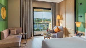 A hotel room with a sofa, armchair, and bed, a small table with a tray of snacks, and a view of a golf course through a large window. - The Aaureum Resort & International Convention Centre (Bhilwara)