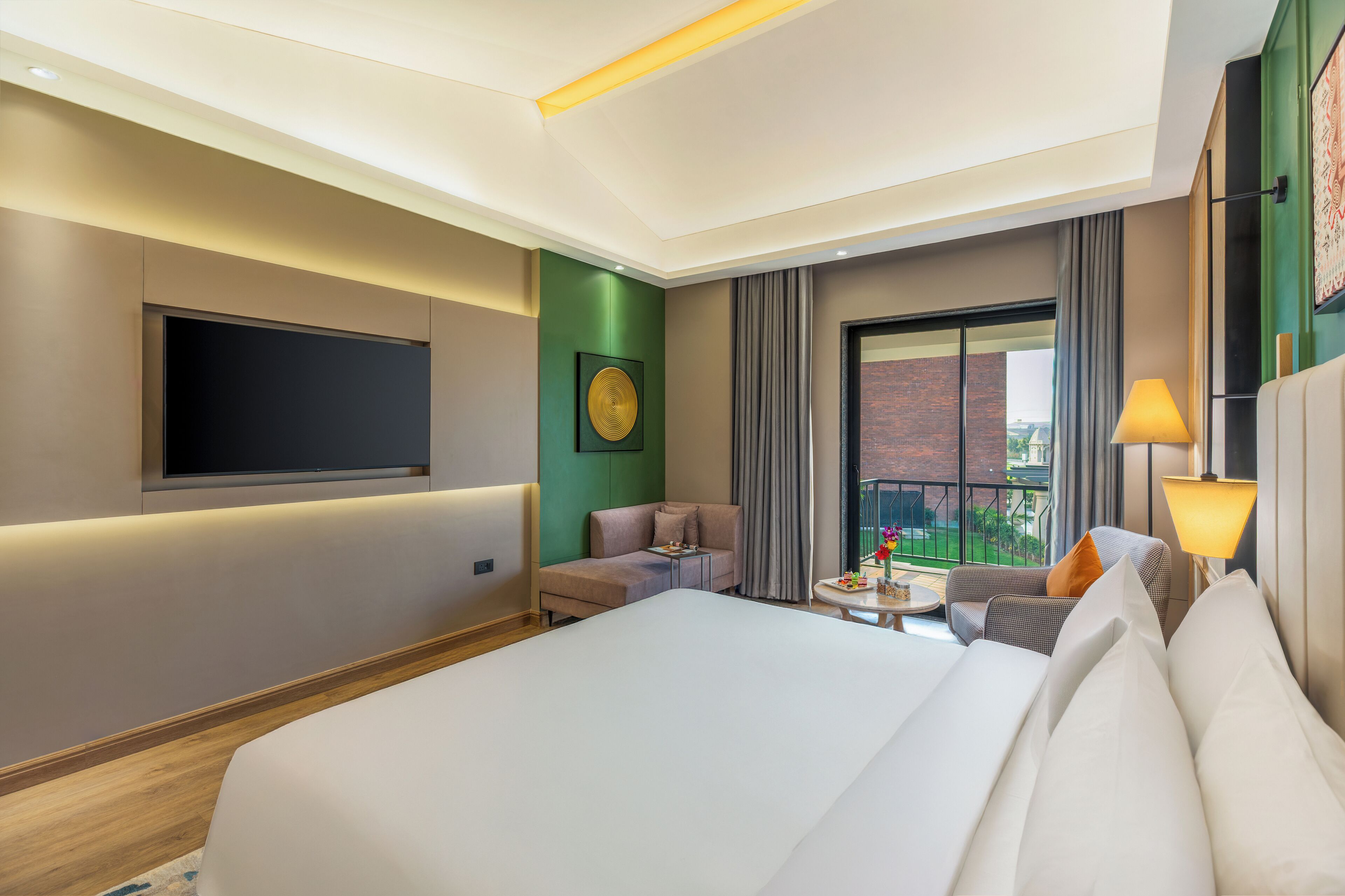 A hotel room with a large bed, a flat-screen TV, a sofa, a small table, and a balcony with a view.