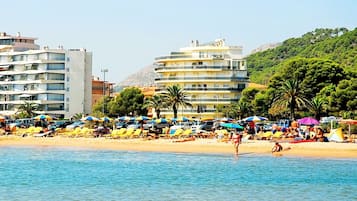 Una playa con tumbonas y sombrillas, un edificio con balcones y montañas al fondo.