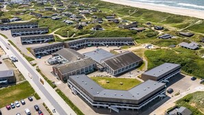 Eine Vogelperspektive zeigt eine Küstenregion mit einem großen Gebäudekomplex, Parkplätzen und einem Strand im Hintergrund.