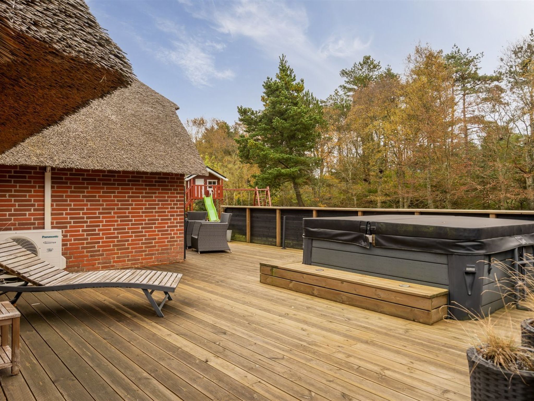 Een houten terras met een gebouw met rieten dak, een jacuzzi en een glijbaan.