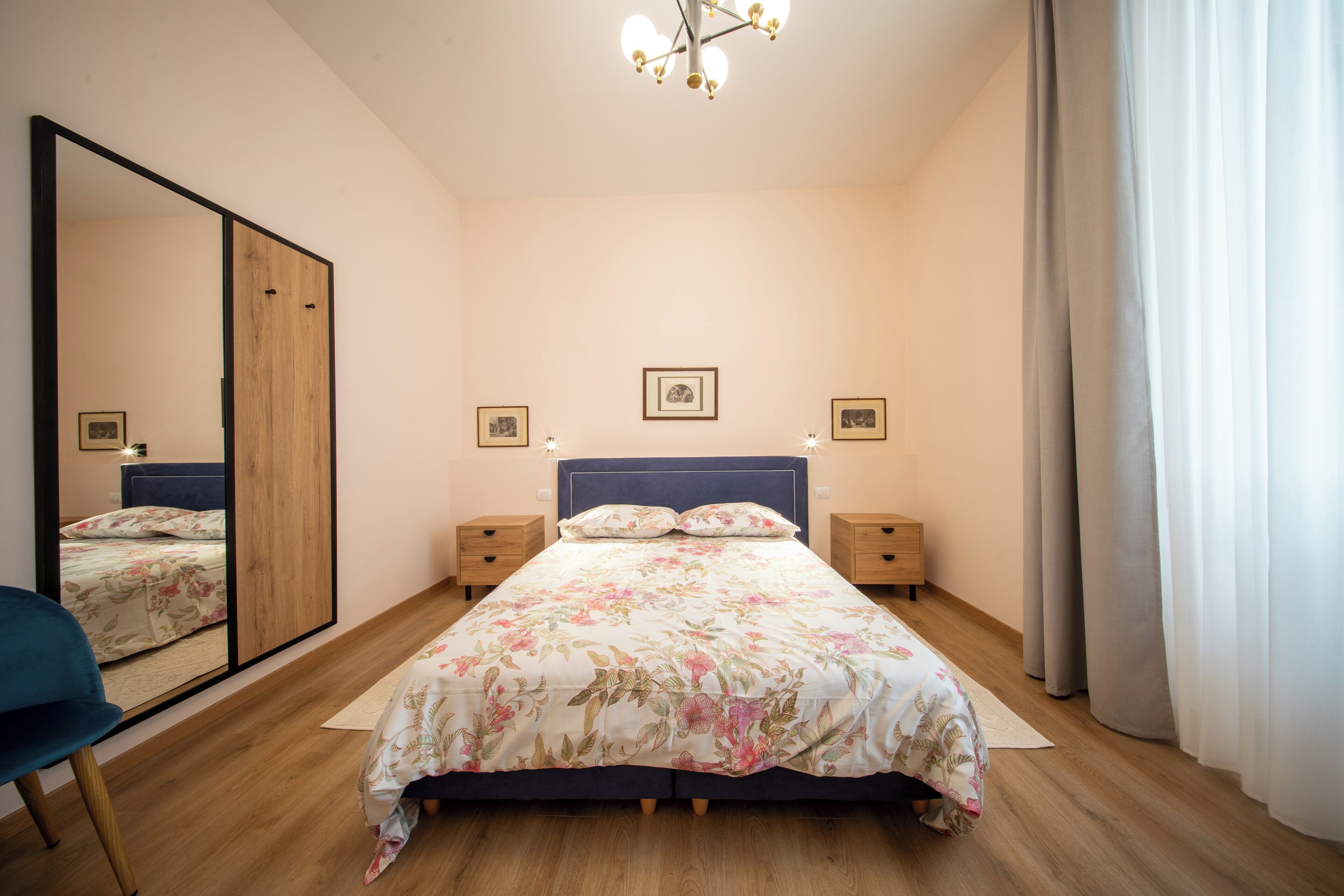 Quarto com um espelho grande, cama com roupa de cama florida, mesas de cabeceira de madeira e luminária no teto.