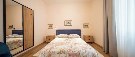 Quarto com um espelho grande, cama com roupa de cama florida, mesas de cabeceira de madeira e luminária no teto.