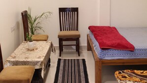 A small, well-lit room with a bed, a table with a plant, a chair, and a framed picture on the wall. - my homestay jaipur soni2700 (Jaipur)