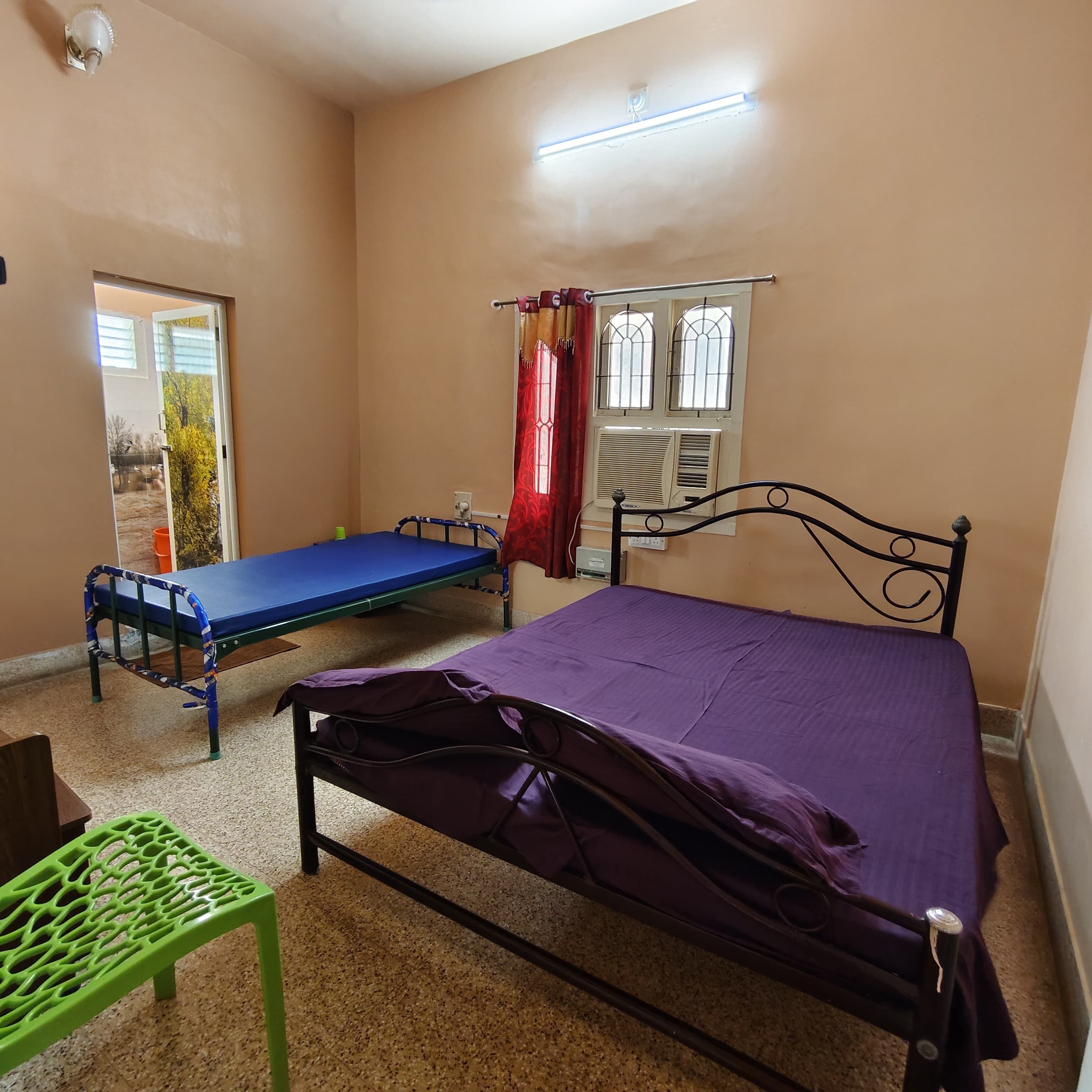 A room with two beds, one with a purple bedspread and another with a blue bedspread. There is a green chair, a window with curtains, and an air conditioning unit on the wall.