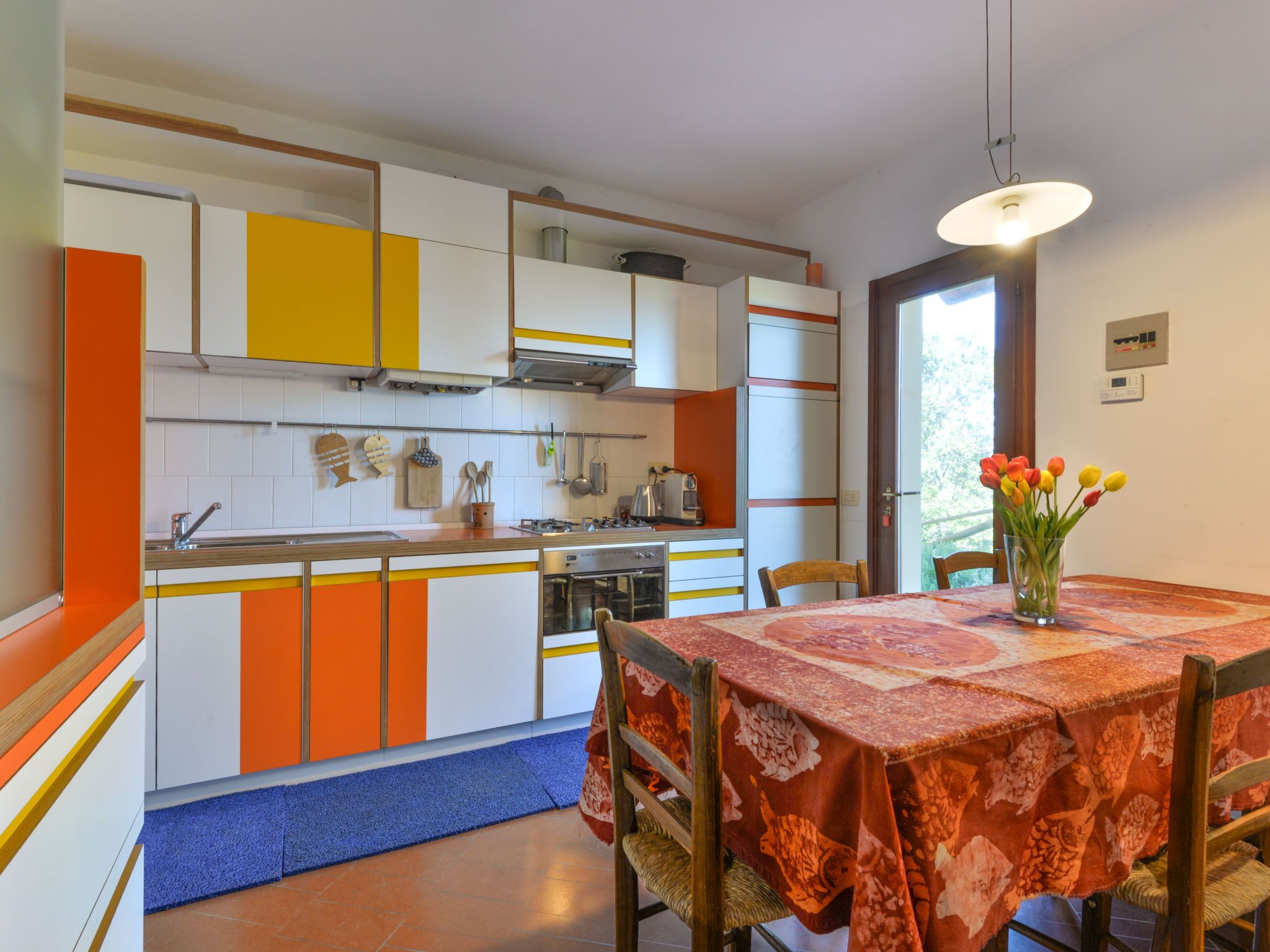 A kitchen with orange and white cabinets, a dining table with a floral tablecloth, and a vase of tulips.