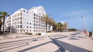 A multi-story hotel with a tiled pavement, palm trees, and a clear sky. - Beachfront T2 Apartment with Sea View and Shared Pool (Quarteira)
