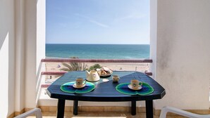 A balcony with a table set for two, overlooking the sea, with a view of the beach and a red and white fence. - Beachfront T2 Apartment with Sea View and Shared Pool (Quarteira)