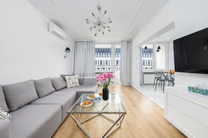 A modern living room with a glass coffee table, grey sofa, and a dining area with a black TV and a table with chairs. - P&O Apartments Boya-Żeleńskiego (Warsaw)