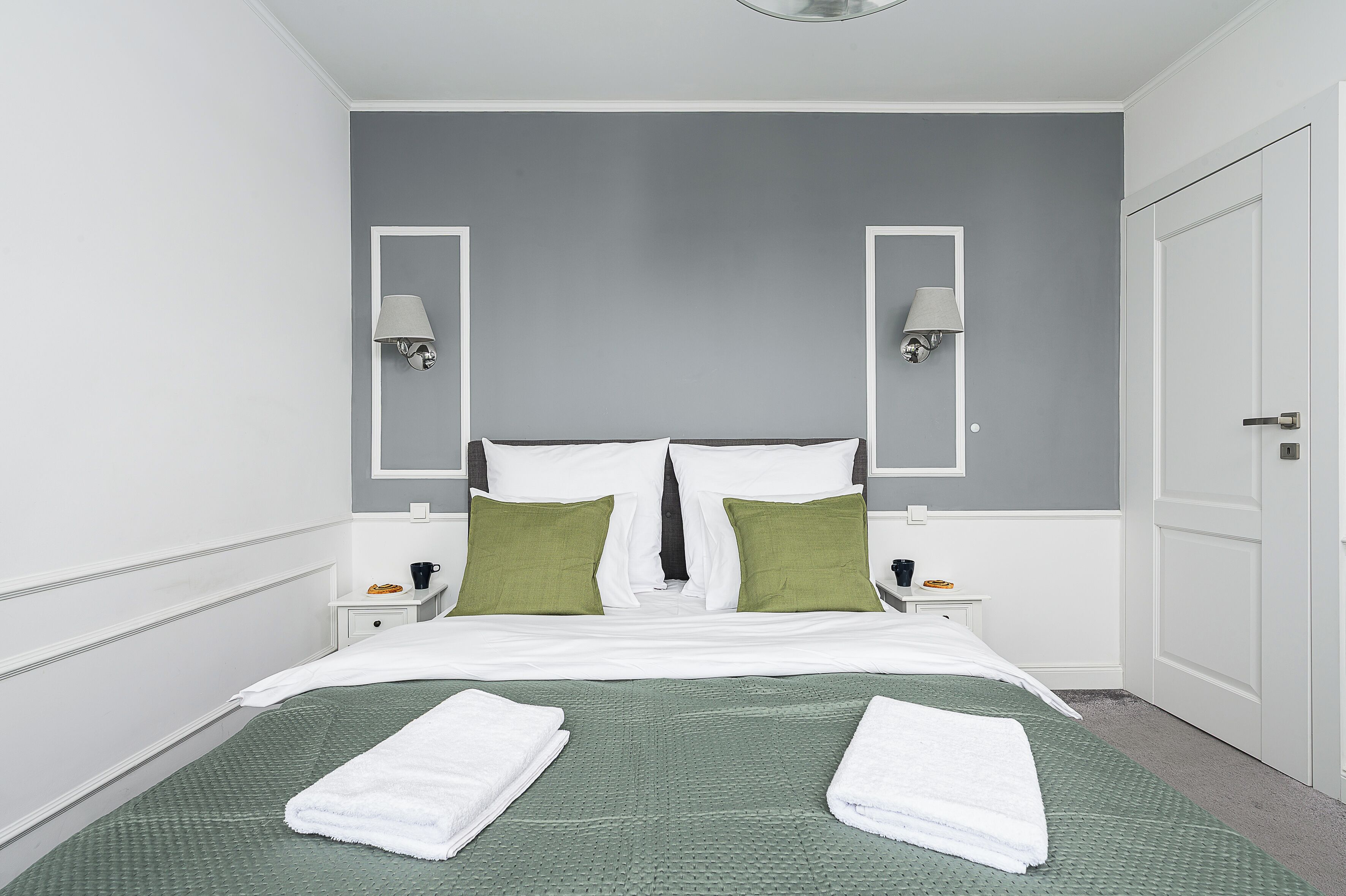 A neatly made bed with white and green bedding, two nightstands with lamps, and a gray wall.