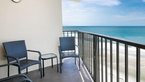 A balcony with two wicker chairs and a small table, overlooking a beach with clear blue water.