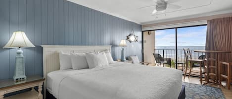 A hotel room with a large bed, a ceiling fan, a balcony with a view of the ocean, and a table and chairs outside.