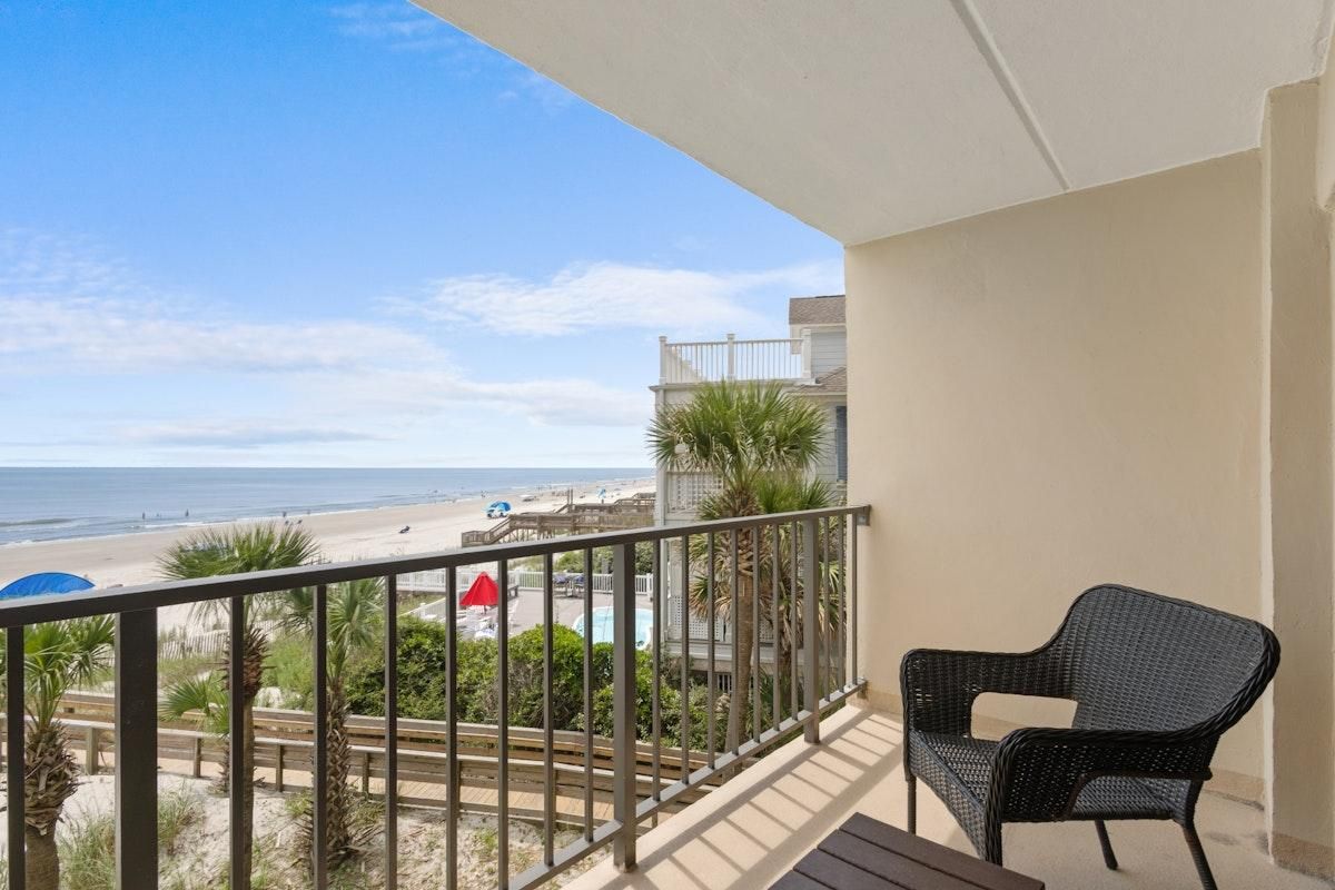 A balcony with a view of the beach, a chair, and a table.