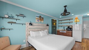 A bedroom with a bed, a chair, a wooden headboard, a small kitchenette, and shelves with various items. - Beaches and Waves Resort View 2nd Floor (Pawleys Island)