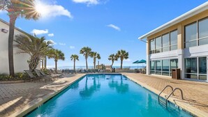 A swimming pool with lounge chairs, palm trees, and a building with large windows. - Beaches and Waves Resort View 2nd Floor (Pawleys Island)