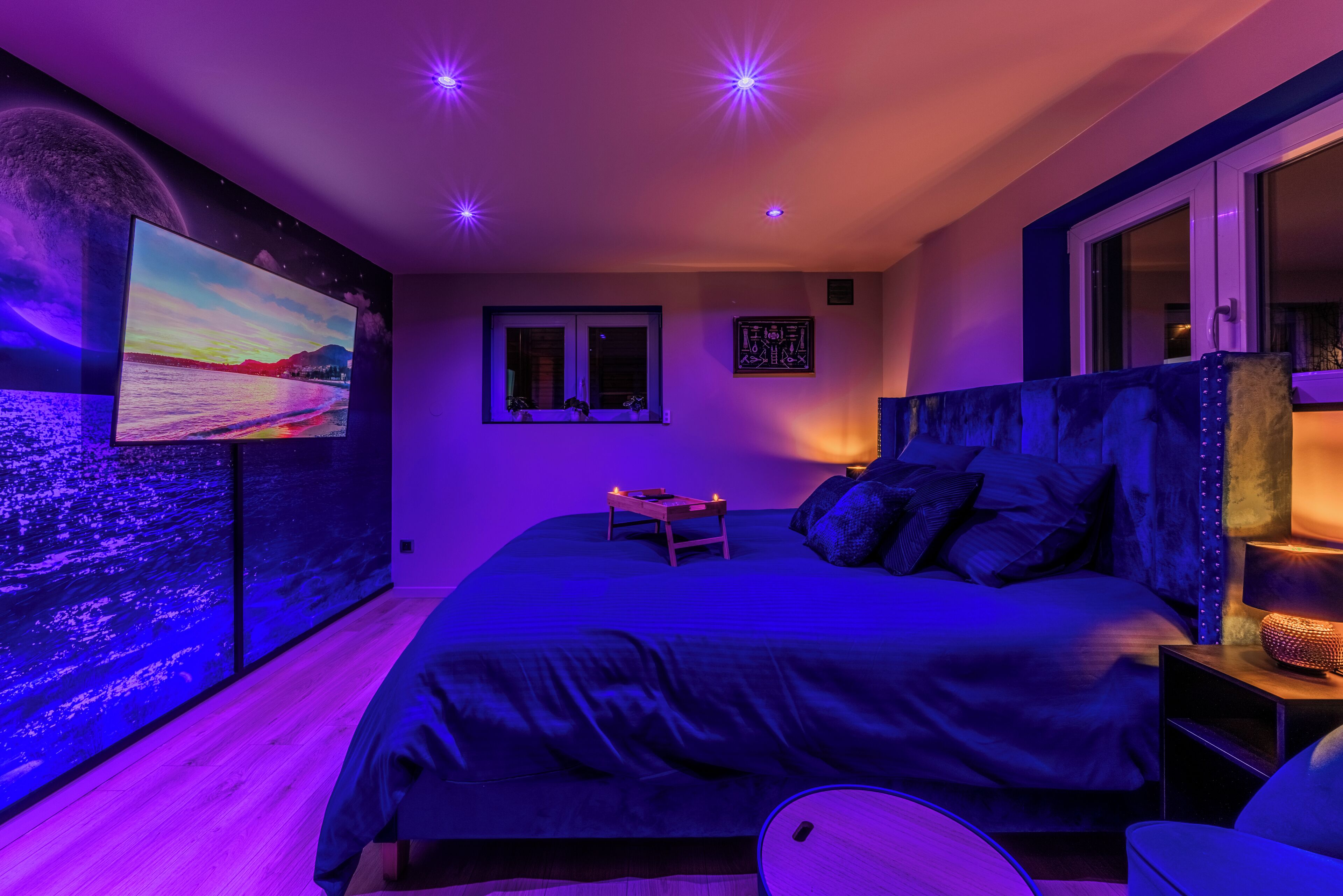 A bedroom with a large bed, a TV mounted on the wall, a window, and a nightstand with a lamp.