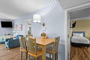 Une pièce comprenant une table à manger, des chaises, un téléviseur fixé au mur, un lit avec une tête de lit et un plancher en bois.