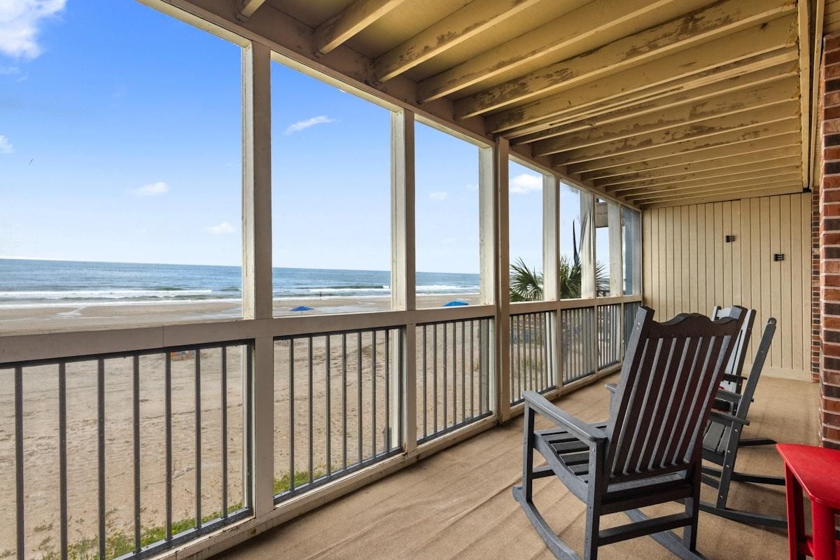 A covered balcony with a view of the beach, wooden chairs, and a red table.